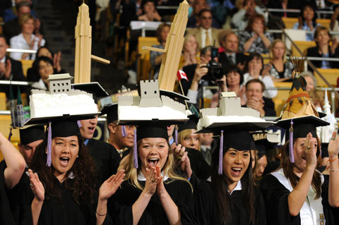 Four female students wearing black gowns and caps. On top of their caps are unique architecture models including a tepee