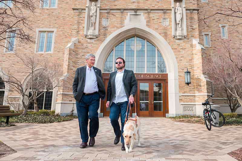 Two men and a seeing eye dog walk in front of the College of Science building on Notre Dame's campus.