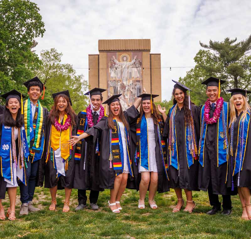 Nine men and women wearing graduation robes and caps smile arm in arm in front of Hesburgh Library at the University of Notre Dame.