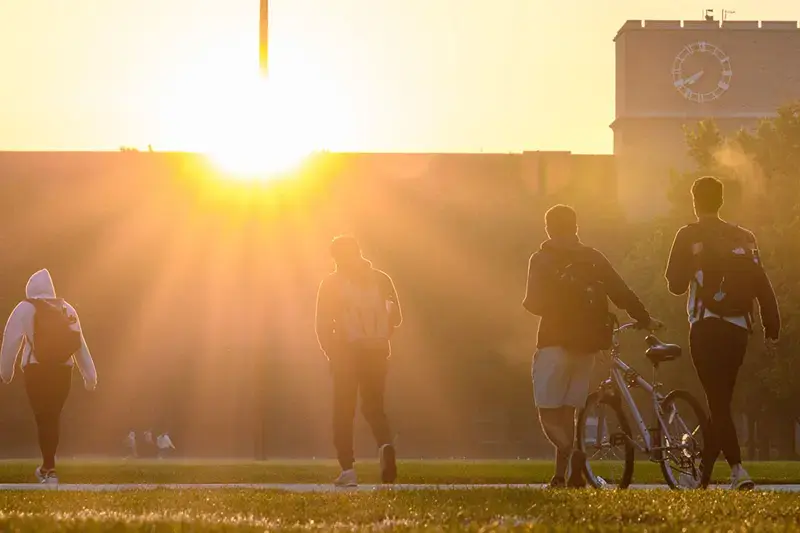 A group of students walk in the direction of an an academic building silhouetted by the sun.