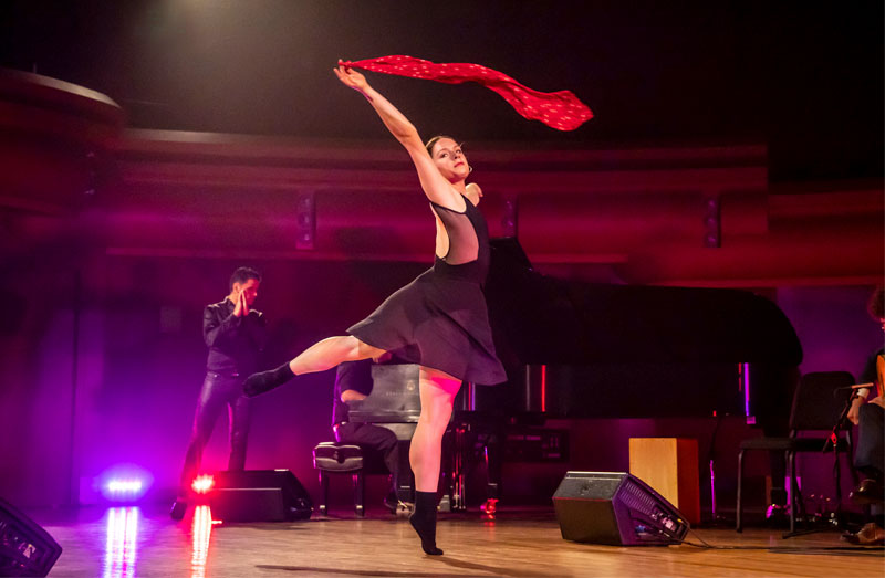 A female dance wearing a black leotard and black mesh skirt stands in an arabesque while holing a red scarf over her head.