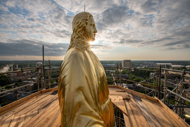A side view of the statue of Mary on top of the Golden Dome. The sun is shining directly behind her.