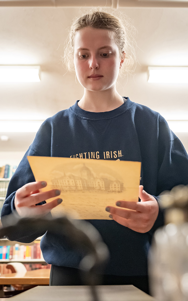 A blonde white female student wearing a dark blue sweatshirt holds a yellowed letter. You can see the front of the letter through the back, showing a gray illustration of the main building at the University of Notre Dame.