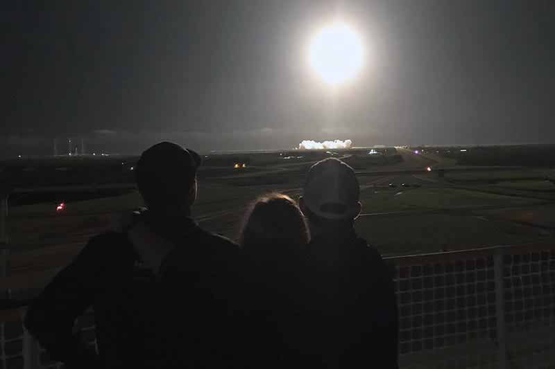 Three people stand closely next to each other and look to the sky at the night sky as a spaceship takes off.