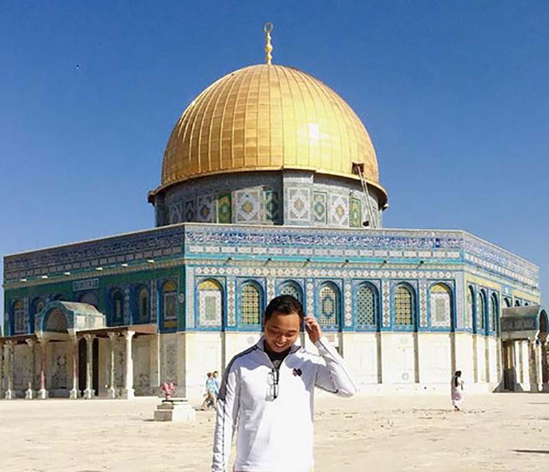 A young man stands in front of a building with a golden dome on a sunny day.