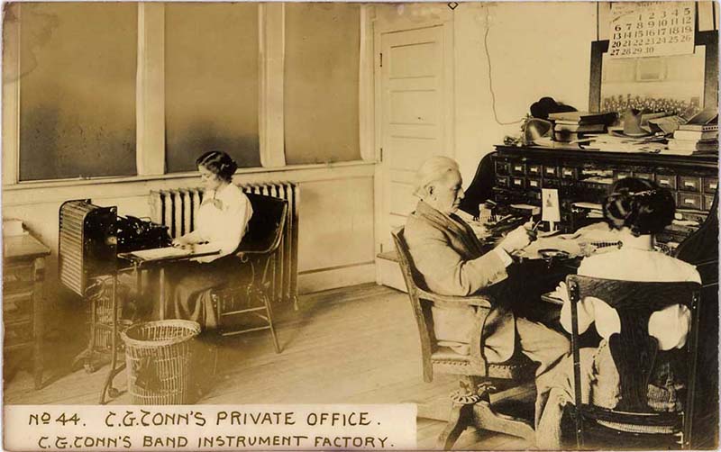 A sepia-toned archive photo of a white haired man sitting at a desk in an office, beside a woman. Another woman sits at a seperate desk with a typewriter.