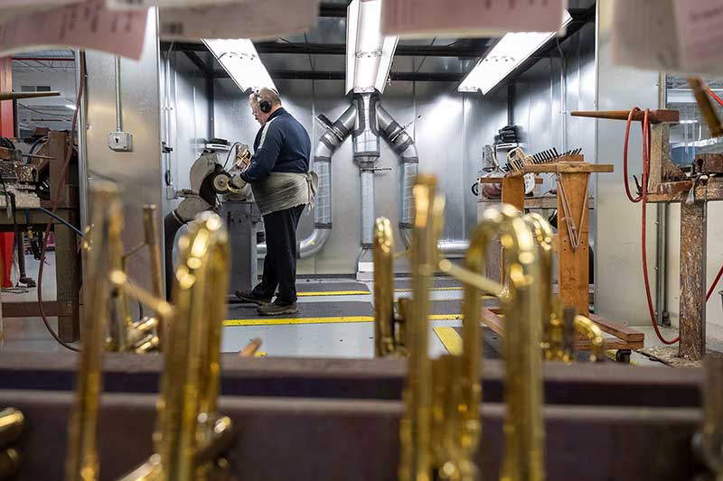 Blurred trumpets in the foreground in a factory room. In the background a white man, in focus, wearing ear protection uses a machine to buff blemishes in the brass.