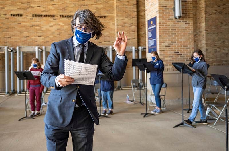 A masked man holding sheet music conducts a group of singers in the background.