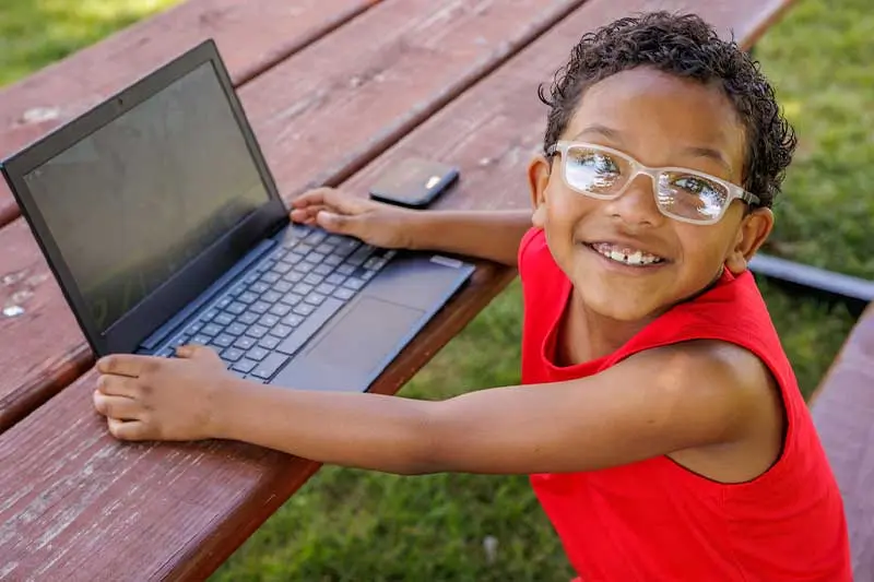 A little boy sits at a picnic table using a laptop. Next to him is a small personal hotspot