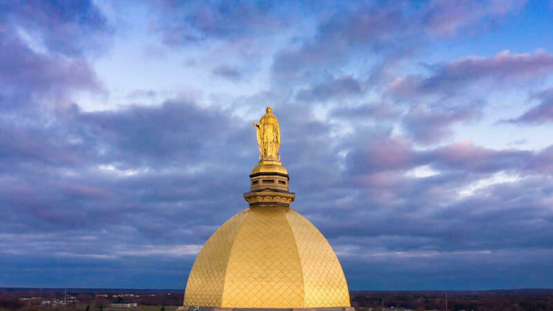 The Golden Dome of the Main Building at the University of Notre Dame, topped with the statue of Mary, against a cloudy blue and purple sunset sky.