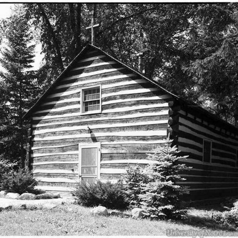 Black and white photo of a small, dark, horizontal-log cabin with white chinking, a cross on the peak of the roof, and surrounded by trees and shrubs.