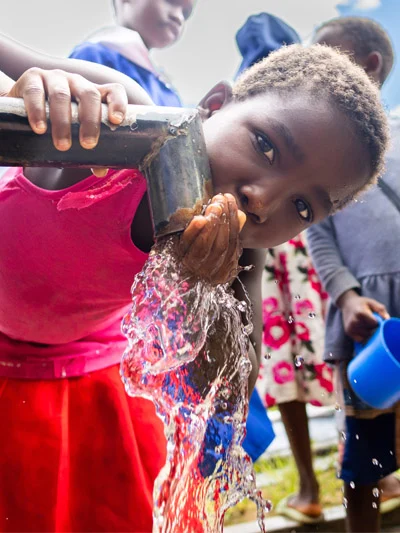 A young child in a pink shirt and red skirt drinks clean water directly flowing from an outdoor hand-operated water pump.