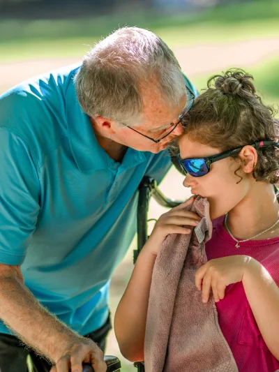 A grandfather kisses the forehead of his granddaughter in a wheelchair wearing sunglasses as they share a quiet, affectionate moment outdoors.