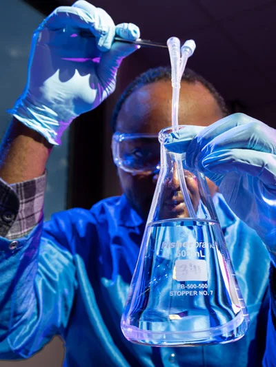 A researcher wearing safety goggles, a lab coat, and blue nitrile gloves uses tweezers to lower tubing into a glass flask filled with clear liquid.