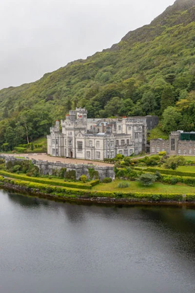 Kylemore Abbey sits at the base of a steep, green mountain along the edge of a calm lake under an overcast sky.
