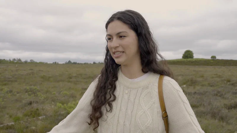 A young woman with long, dark curly hair wearing a cream cable-knit sweater stands in an open bog landscape under an overcast sky.