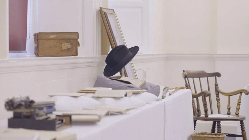 AA table covered in a white cloth displays historical items including open books on pillows, a black fedora leaning against a framed picture, and a wooden chair nearby.