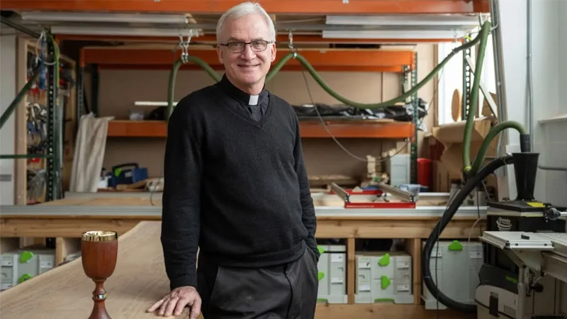 A Catholic priest wearing a black sweater, clerical collar, and glasses standing in a woodworking workshop near a wooden chalice.
