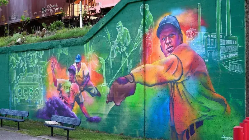 A vibrant mural on a green concrete wall depicts baseball players, industrial scenes, and a figure pointing, with a train car visible above.