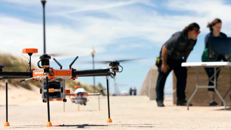 An orange and black drone sits on paved ground outdoors with two operators in the background monitoring data on a computer.