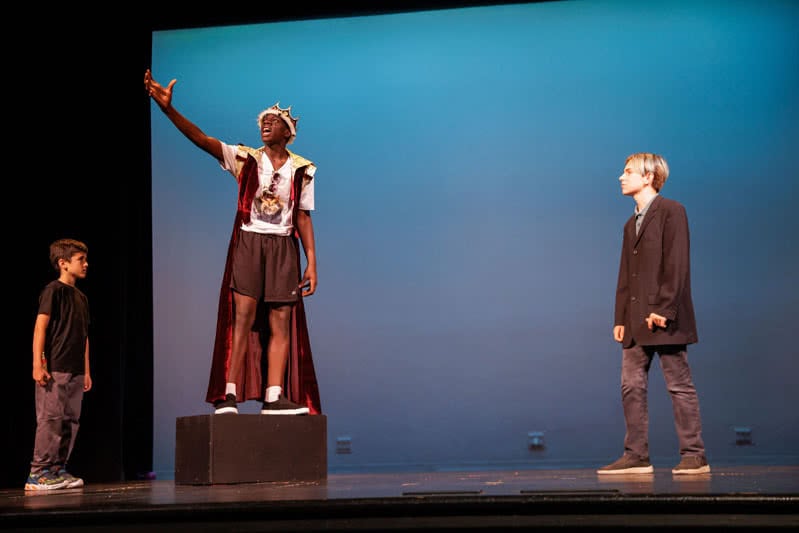Students on stage performing Shakespeare. One student is standing on a box with his hand in the air and crown on his head.