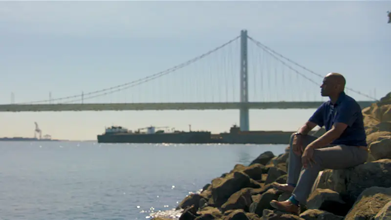 Ernest Morrell sits on some rocks overlooking a river and a bridge.