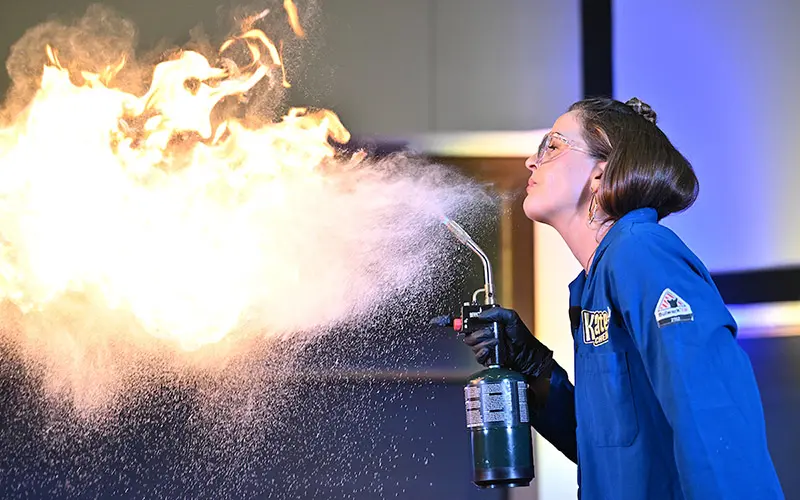 A woman in a blue lab coat, safety glasses, and black gloves aims a small torch at a burst of flames, demonstrating a chemical reaction.  Fine particulate matter hangs in the air around the flame.