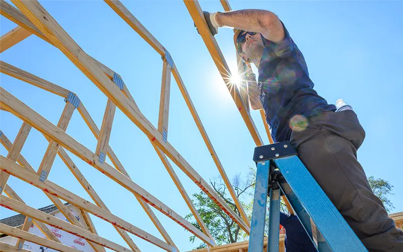 A white male wearing safety glasses stands on a blue stepladder, securing wooden roof beams against a bright blue sky. The sun flares behind him, highlighting the framework of the house under construction.