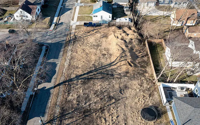 Aerial view of a vacant, dirt-covered lot surrounded by single-family homes. Barren trees cast long shadows across the lot. It is late winter.