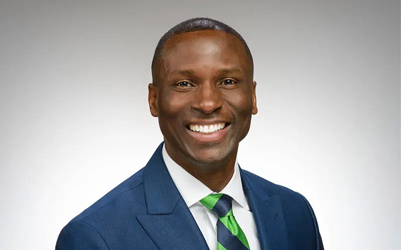 A black man in a blue suit coat and green and blue wide striped tie.