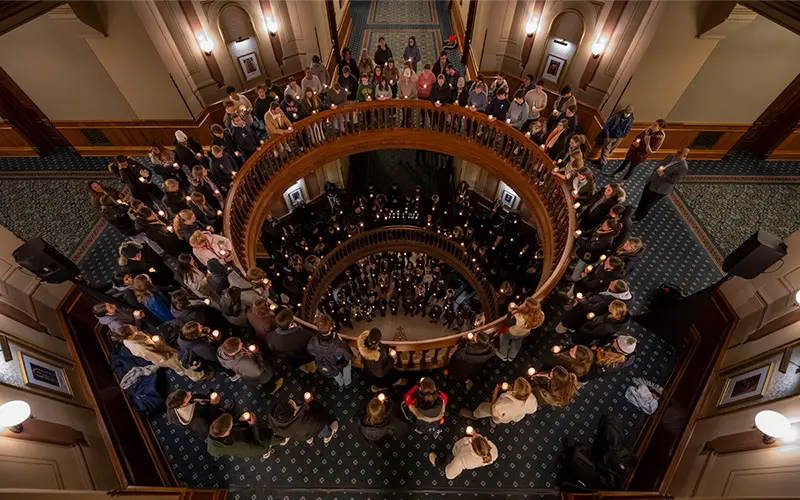 A high-angle view of a candlelight vigil held within a grand, spiraling staircase. Students line the wooden railings on multiple levels, each holding a lit candle, creating a warm, glowing atmosphere. The ornate details of the building's interior are visible in the soft light.