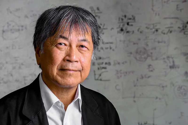 Headshot of a Asian man with graying hair and a dark blazer, standing in front of a whiteboard filled with scientific equations and diagrams.