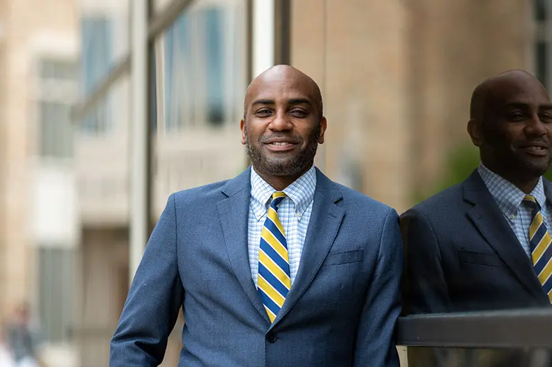 A bald black man wearing a navy blue suit and a blue and gold striped tie smiles as he leans against a glass wall reflecting a blurred image of himself on the Notre Dame campus.