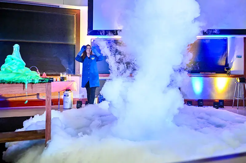 A white female scientist in a blue lab coat reacts to a large white cloud of foam expanding across a classroom floor.  Bright green slime spills over a table to the left. A chalkboard and projection screen are visible in the background. Colorful lights illuminate the scene.