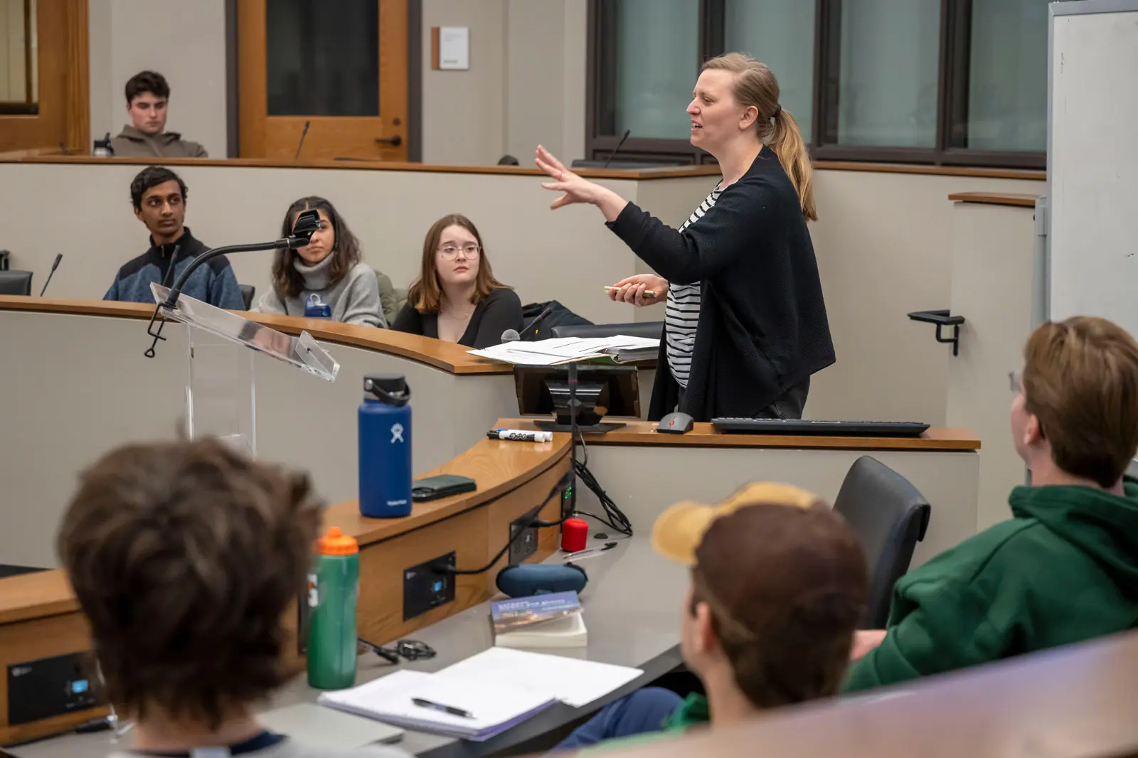 A white female professor gestures while lecturing to students in a tiered lecture hall. The students sit at a curved desk facing the professor, who stands with papers at a podium.