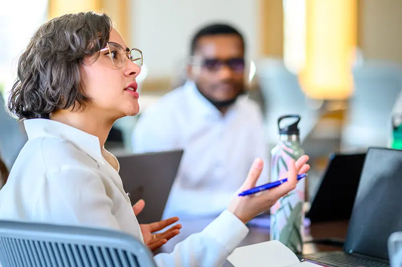 A white female wearing glasses and a white shirt gestures with a pen in hand while speaking during a group discussion. A student is slightly out of focus in the background. Laptops and a water bottle rest on the table.