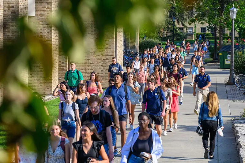 A large group of Notre Dame students walk on a campus sidewalk between brick buildings. Green grass and trees line the walkway.