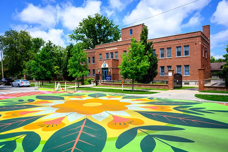 A vibrant street mural featuring a large, stylized flower with orange petals and green leaves dominates the foreground. In the background, a red brick building, possibly part of the University of Notre Dame, stands on a tree-lined street under a blue sky with fluffy white clouds.