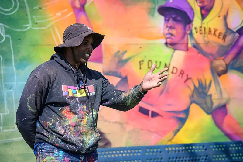 An artist wearing a paint-splattered black hoodie and bucket hat gestures toward a vibrant mural. The mural depicts young baseball players, one with a jersey that reads Foundry.