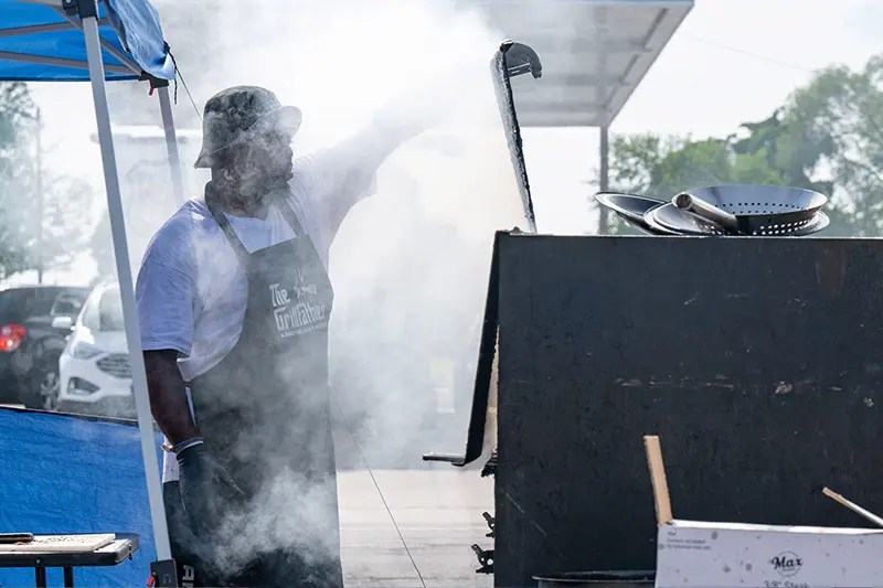 A black man wearing a black apron that reads “The Grillfather“ tends to a large, smoking grill.  Smoke billows around them and cooking utensils rest on top of the grill. A blue pop-up tent is partially visible on the left.