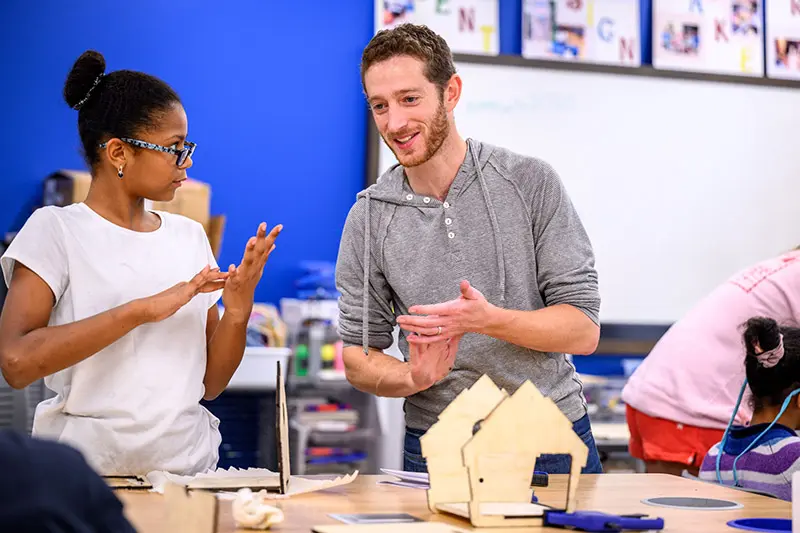 A young black girl and a white man gesture with their hands while discussing small wooden houses on a table in a classroom. Other children work on projects in the background.