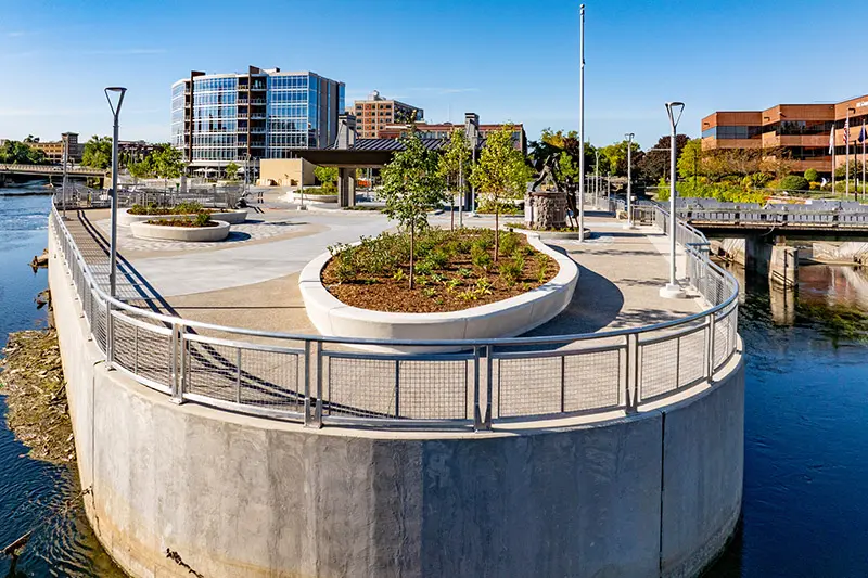 A curved walkway extends into a river, featuring a concrete retaining wall, metal railings with mesh, and landscaped planters. Modern buildings and a bridge are visible in the background under a clear blue sky.