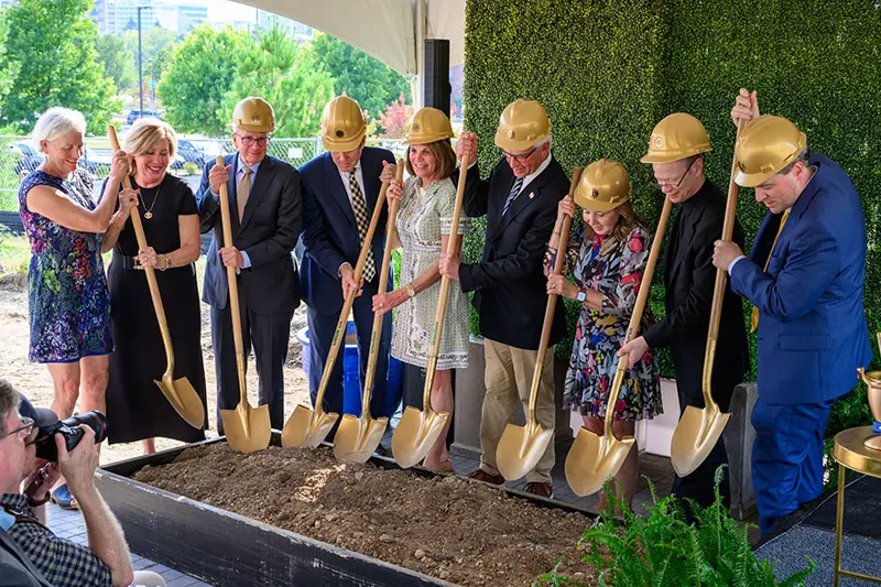 A group of people wearing gold hard hats ceremoniously use gold shovels to break ground on a construction project. A photographer captures the moment. A green wall of foliage provides a backdrop.