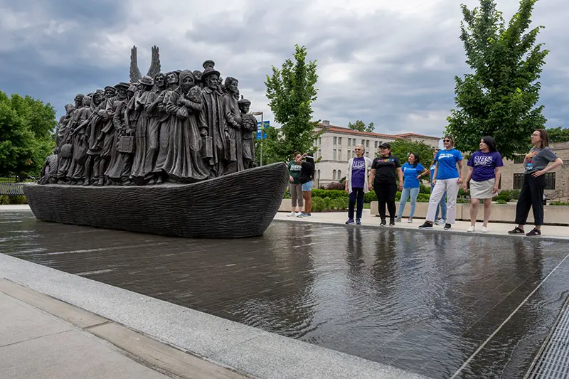 A group of people stand near a large bronze sculpture of immigrants on a boat, symbolizing the journey to a new life.  A partially cloudy sky is above.