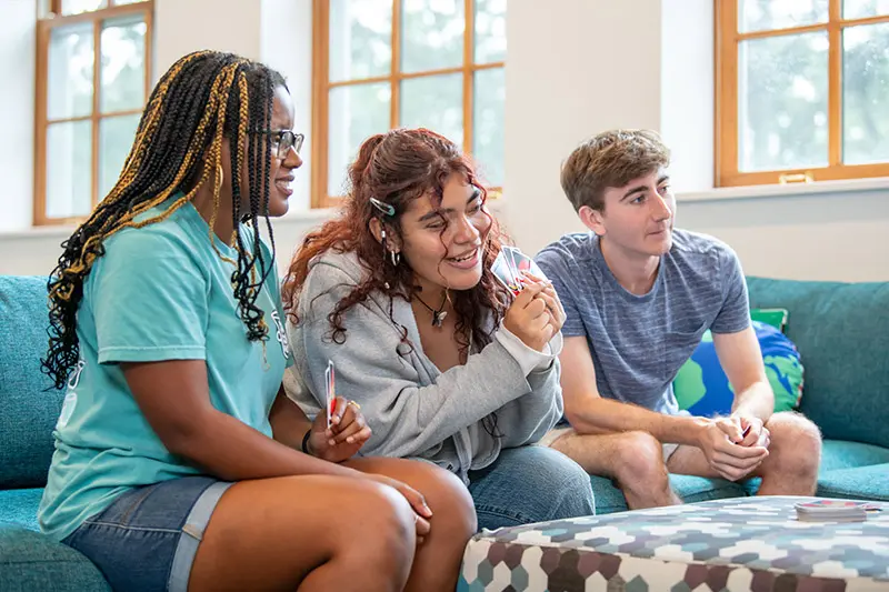 A black women, latino woman, and white man sit on a teal couch playing cards in a well-lit room with large windows. A geometric-patterned coffee table sits in front of them with cards laid upon it.