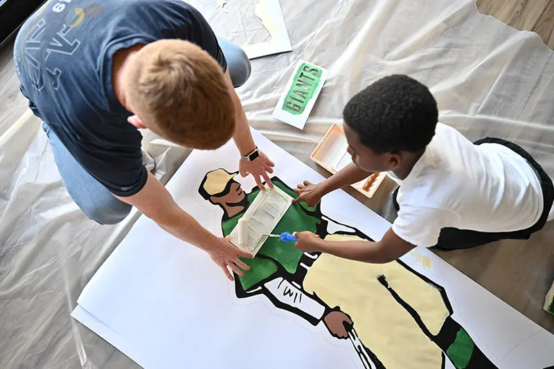 A black boy and a white man kneel over a large piece of paper on a drop cloth, carefully painting a life-sized illustration of a baseball player wearing a green shirt and khaki pants.  The boy uses a small paint roller to apply tan paint.  A small green and white sign reading GIANTS lies nearby.