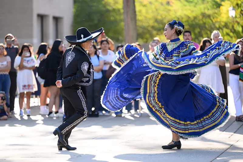 A man and a woman perform a traditional Mexican folk dance outdoors. The female dancer twirls in a vibrant blue dress with gold and green accents, its full skirt billowing around her. The male dancer, wearing a black charro suit with silver embroidery and a wide-brimmed sombrero, faces her. A small crowd watches in the background.