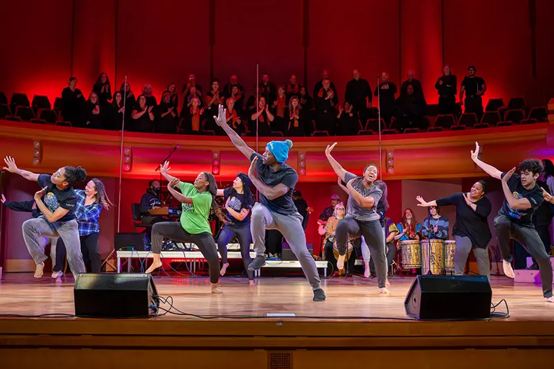 Dancers perform on a brightly lit stage with a choir and band in the background. The dancers extend their arms and legs in various poses, conveying energy and emotion. The red and gold hues of the auditorium enhance the vibrant atmosphere of the performance.