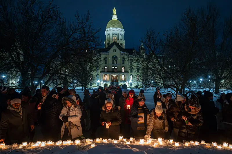 At night, a crowd of people gathers in the snow in front of the Main Building at the University of Notre Dame. They hold lit candles, creating a warm glow against the cold, snowy ground and the dark silhouette of the iconic golden dome.