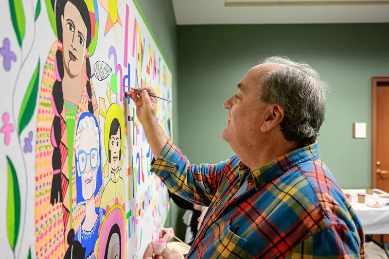 A man in a plaid shirt paints a colorful mural depicting diverse faces and the words My Sky and Heart at the Family. He holds a paintbrush and small container of pink paint.